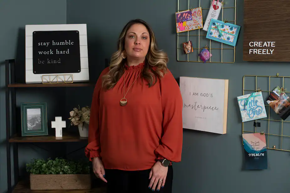 The designer, wearing rather frumpy attire, stands in her office, which is adorned with pithy sayings and religious paraphernalia